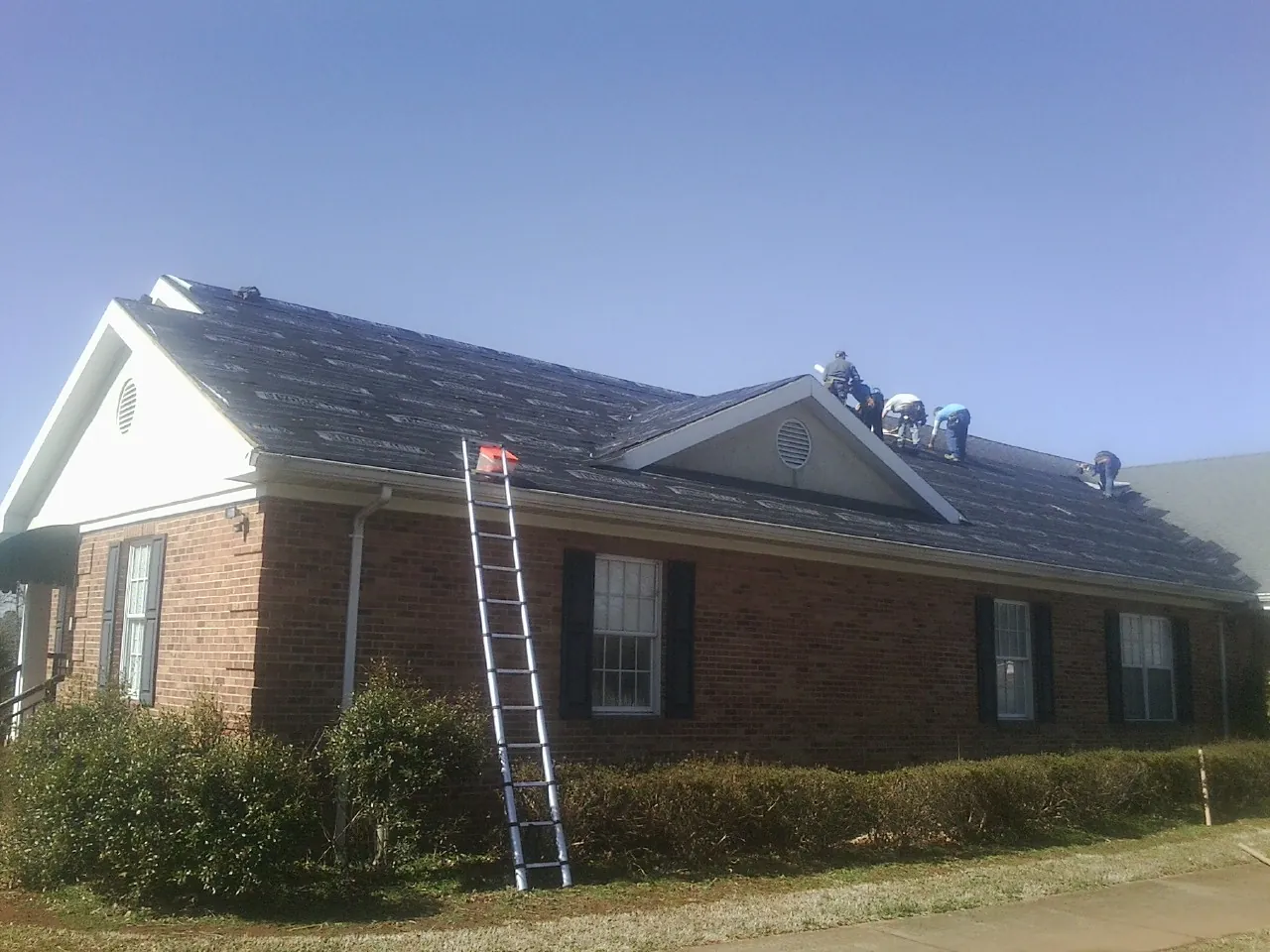 Crew tearing shingles off a brick ranch roof with a ladder set — Emergency Roof Repair work by our crew in Lenexa