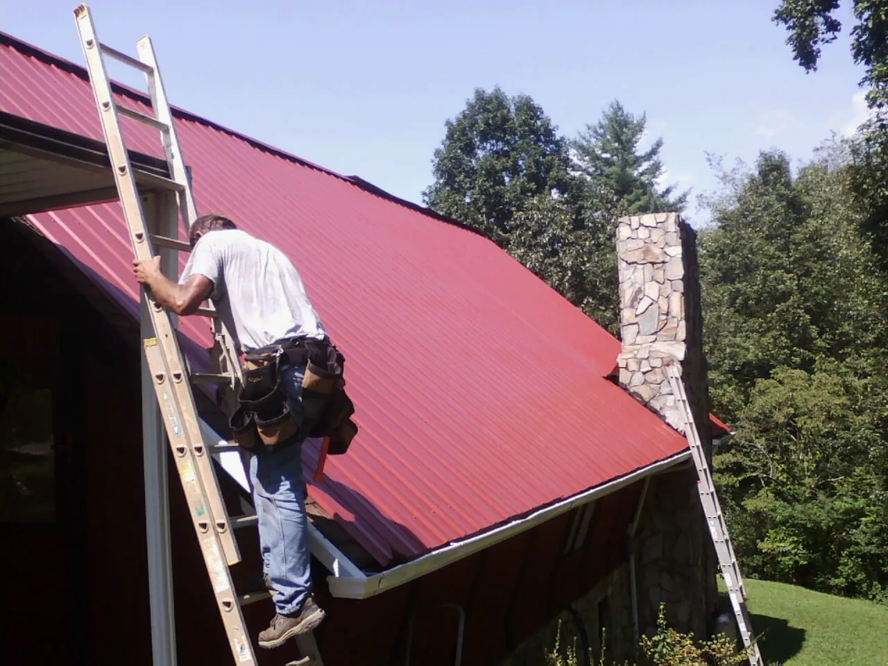 Roofer on a ladder beside a red metal roof and stone chimney — Soffit & Fascia Repair by Roofing Brothers in Lenexa